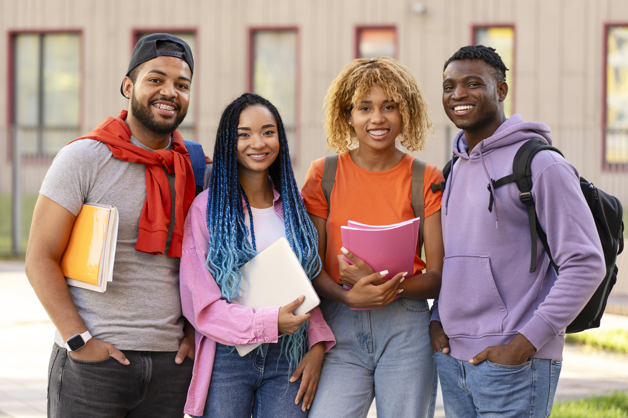 Smiling stylish African American student with backpacks, holding books standing in university campus. Portrait of happy friends looking at camera on the street. Education concept